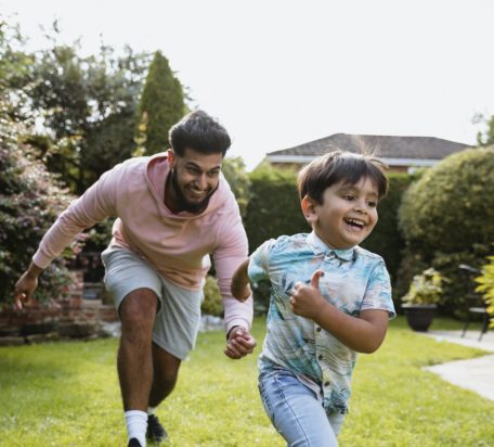 一个年轻人和他的儿子在外面的花园里玩追逐游戏，两人都面带微笑。