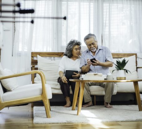 一位年长的男性低头看手机，而他的年长的妻子都坐在客厅的沙发上。