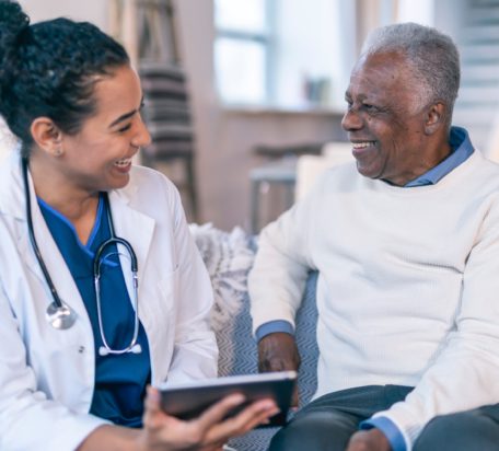 一位女医生拿着一台电子平板电脑，看着一位年长的男病人，两人都坐在病人家中的沙发上微笑。
