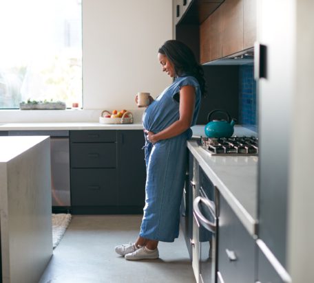 一位孕妇站在家里的厨房里微笑着，手里拿着热饮，低头看着自己的肚子。