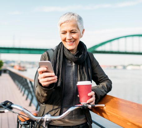 一位年长的女士站在她的自行车旁边，站在栅栏旁，一边微笑着在电话里打字，一边在河边喝咖啡。