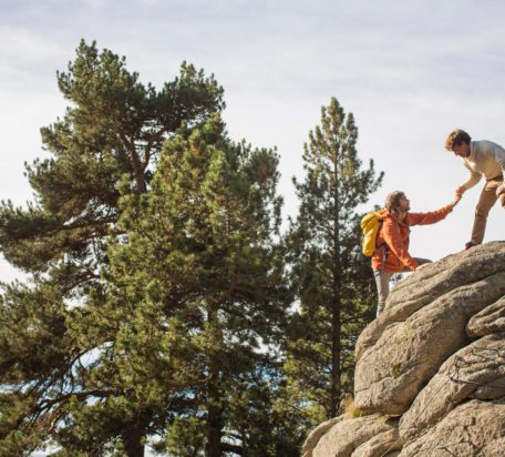 一只雄性在帮助另一只背着背包、穿着橙色夹克的雄性爬上背景有松树的岩石。